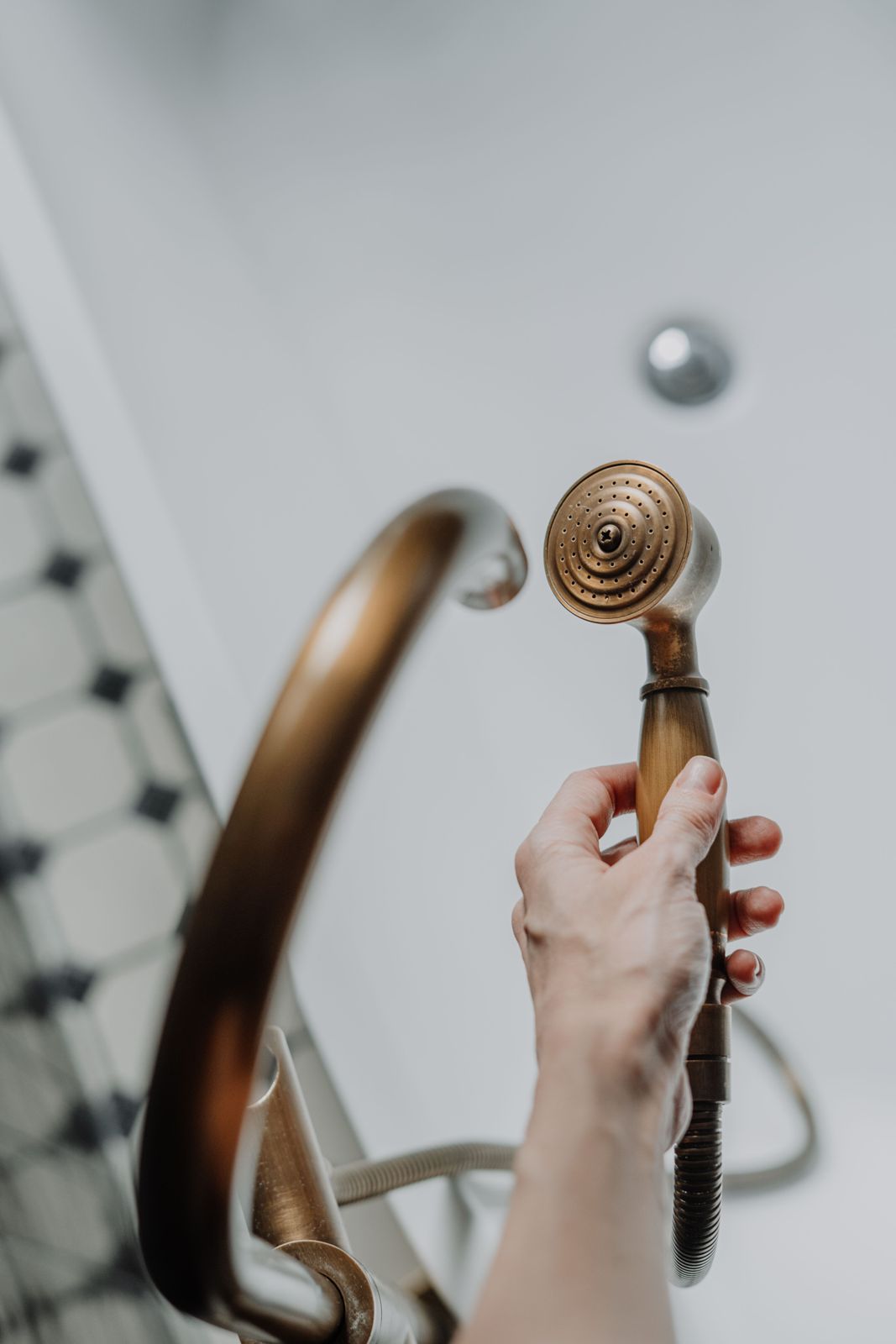 Hand holding an antique brass handheld shower head beside a curved brass bath spout