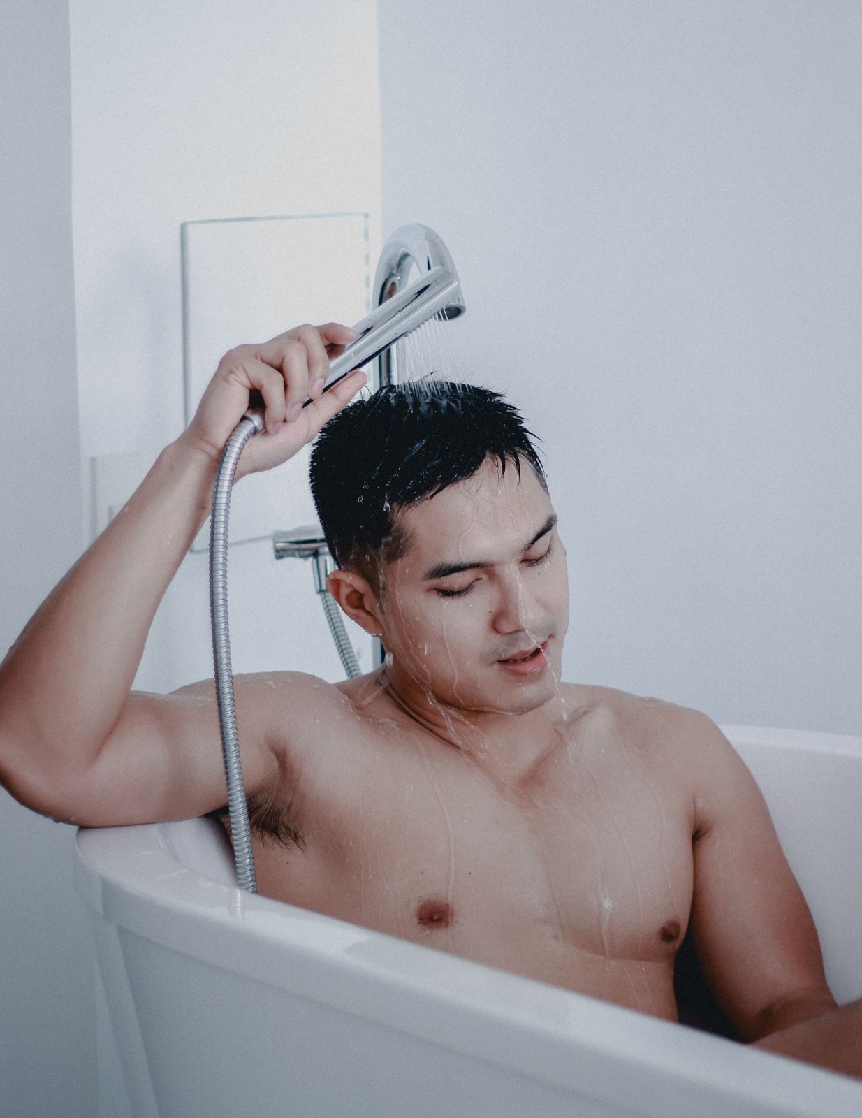 Man sitting in a white bathtub rinsing his hair with a chrome handheld shower
