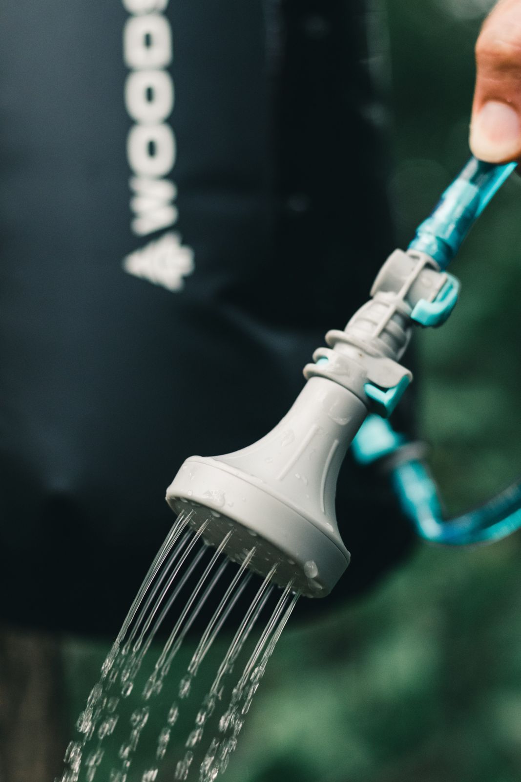 Grey camp shower head with blue tubing spraying water outdoors in front of a dark bag