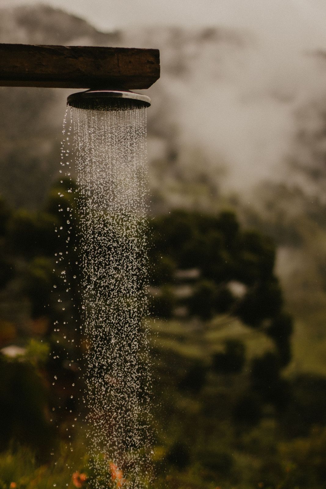 Round shower head fixed under a wooden beam pouring water outdoors with misty green hills behind